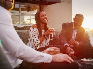 A group of people handing out on a roof top bar sitting down in a conversation pit laughing with drinks during sunset.