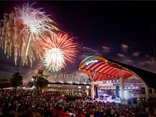 A skyline of downtown Little Rock at night showing the colorful bridge lights, boats in the water, and fireworks in the sky.