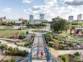 Aerial view of the Riverfront park with people walking, cycling, and playing around the sculptures with the view of downtown Little Rock in the background.